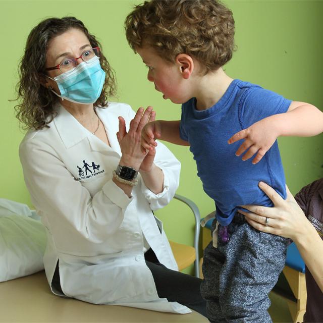 A toddler with curly hair in a blue shirt lifts his arms as he is held by a physical therapist, as Stacy Suskauer look on. 